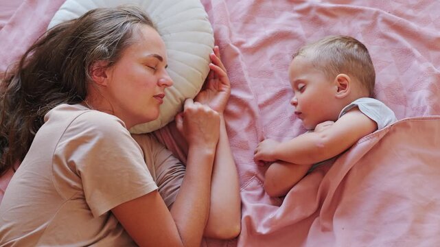 Peaceful mother and her little toddler son sleeping together side by side in cozy bed on pink bedding during quiet daytime nap
