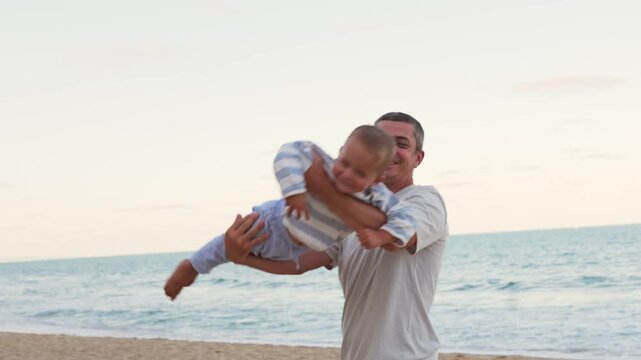 Joyful father lifting and flying his little toddler son in air on sandy sea beach having fun during family summer vacation