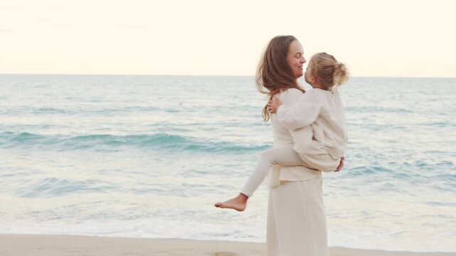 Happy young mother carrying her cute little daughter in arms walking along sandy sea beach during family sunset trip