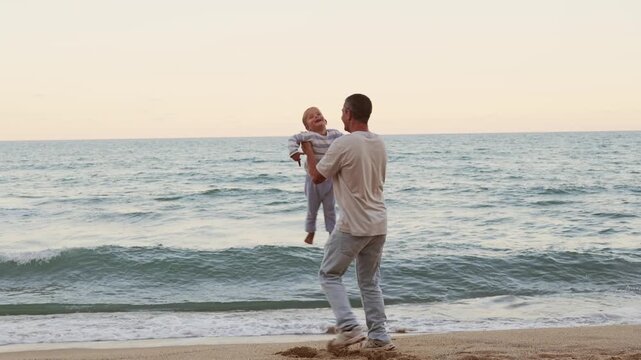 Happy laughing father lifting high his cute little son in arms on sandy sea beach having fun together
