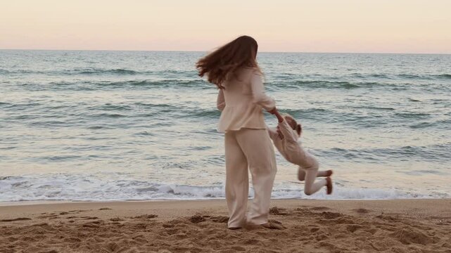 Joyful mother spinning her little daughter in arms while playing on sandy sea beach with older daughter watching