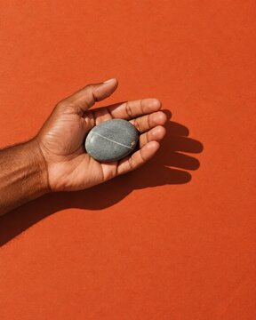 Open male hand with dark skin holding a smooth grey river stone over a matte terracotta background for mindfulness and mental health
