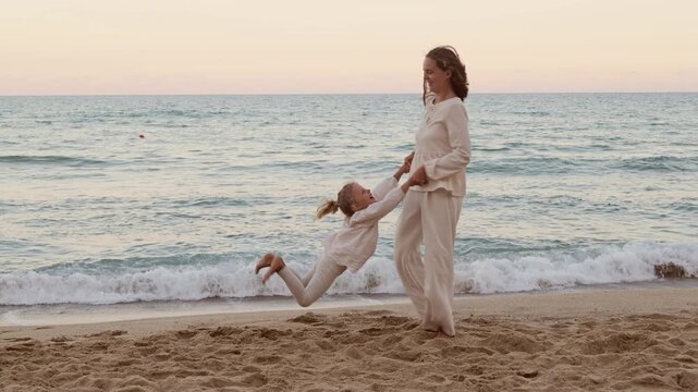 Joyful mother spinning her cute little daughter around in arms while playing on sandy sea beach at sunset
