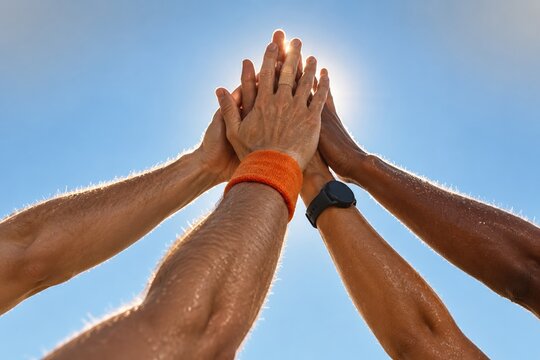 Four arms joining hands in a high five, sweaty skin and diverse skin tones against a bright blue sky for fitness and teamwork concepts