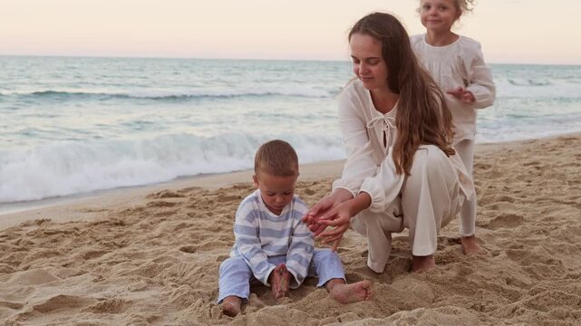 Mother and her two small children sitting on sandy beach and playing with sand together near sea shore