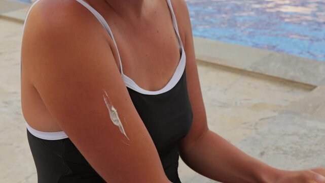 Woman applying sunscreen lotion on her arm sitting near swimming pool during summer vacation