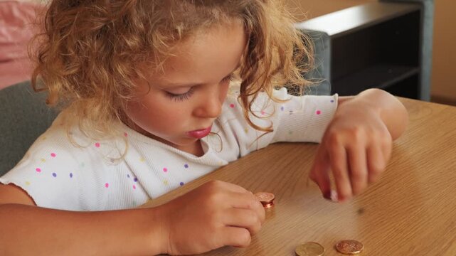 Little girl with curly hair carefully arranging and counting coins on wooden desk at home