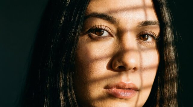 Close-up portrait of a young woman with dark hair, her face partially illuminated by sunlight filtering through blinds, creating striking shadow patterns.