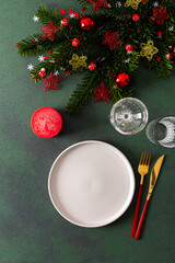Fototapeta premium Top view of a holiday table, showcasing an empty plate, cutlery, and candle on a deep green background. Red tree ornaments and spruse branches enhance the festive ambiance