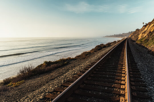 A scenic train track runs parallel to a coastline, with the ocean on one side and a cliff on the other, captured during a soft morning sunrise.