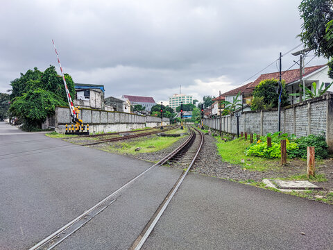 Railway tracks crossing a quiet asphalt road in central Bogor, running between a concrete fence and grassy verge, with trees, rooftops and an overcast sky stretching across the urban skyline.