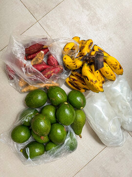Fresh avocados, ripe bananas, red sweet potatoes and bagged coconut water in plastic bags on a white tiled floor, a haul of wholesome real food groceries for healthy family meals.