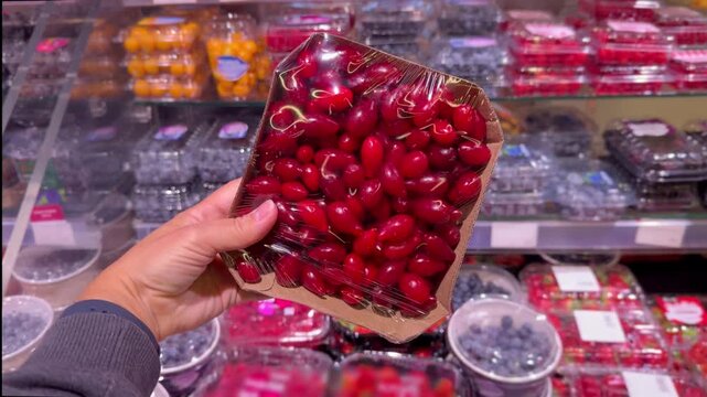 Hand Holding Packaged Berries in Supermarket &ndash; Raspberries, Blueberries, Strawberries and Cornelian Cherries Against Refrigerated Berry Display