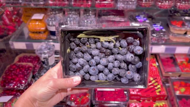 Hand Holding Packaged Berries in Supermarket &ndash; Raspberries, Blueberries, Strawberries and Cornelian Cherries Against Refrigerated Berry Display