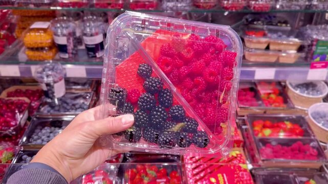 Hand Holding Packaged Berries in Supermarket &ndash; Raspberries, Blueberries, Strawberries and Cornelian Cherries Against Refrigerated Berry Display