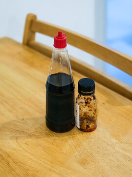 Bottle of dark sweet soy sauce with a red flip cap beside a small jar of chili oil nearly empty, standing on a light wooden table with a wooden chair softly blurred behind in a cafe setting.