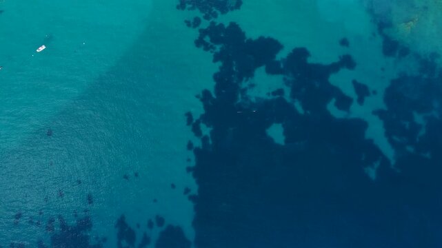 Drone view of turquoise and deep blue ocean water from above. Rocky seabed and seaweed formations visible beneath the clear surface. Stunning contrast between shallow turquoise shallows and deep blue 