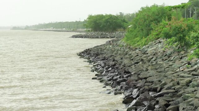 A coastal revetment built with dark riprap stones lines the edge of a murky body of water under an overcast sky in Mosquito Creek, Trinidad, stabilizing the shoreline against erosion