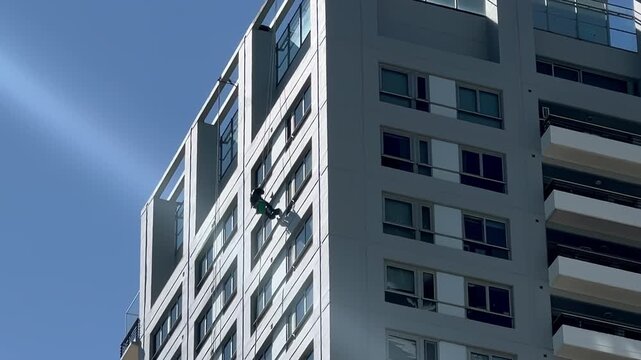 Rope-access window cleaner descending modern apartment building facade with harness safety equipment. Argentina, Buenos Aires