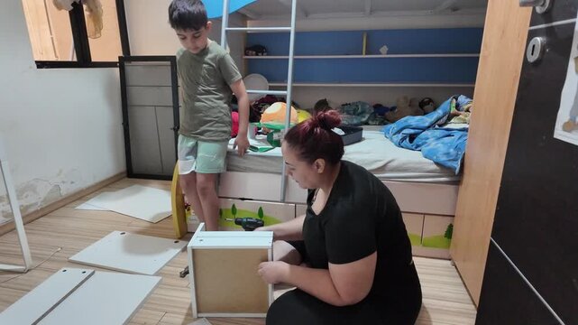Young woman sits floor, assembling storage unit. Child watches nearby, standing beside bunk bed. Room has wooden floors, scattered packing materials. Both appear focused. Perfect for family, DIY