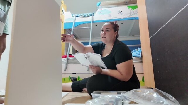 Woman reads manual while assembling furniture. Child plays nearby on bunk bed. Room shows partial setup. She appears focused, calm. Great for home improvement or parenting visuals