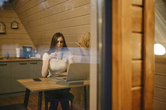 View from outside at a woman sitting at a table with cup of tea with laptop in log cabin in the evening
