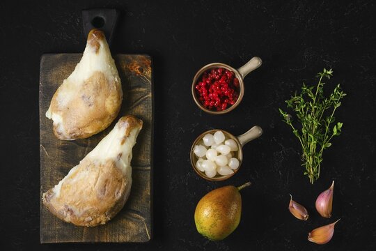Two large duck confit covered with congealed fat on wooden board next to pear, lingonberry, garlic and thyme on dark background in moody lifestyle setting
