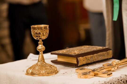Golden chalice, ornate holy book, and decorative cross resting on white tablecloth in a church setting