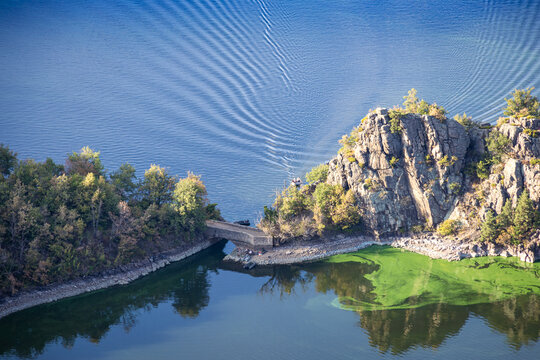 Rocky cliff edge overlooks the calm blue waters of the Loire river at Plateau de la Danse. Rugged granite formations frame a tranquil reservoir landscape near Saint Victor sur Loire.