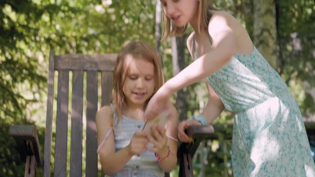 Absorbed girl looking at smartphone screen on wooden chair gets interrupted as older sister snatches phone away playfully, capturing child screen addiction struggle during outdoor play.