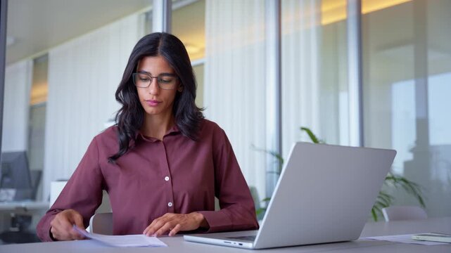 Serious middle eastern business woman in 30s checking paperwork and documents at office desk, indian analyst comparing data with laptop and typing report, medium shot of accountant at daily work.