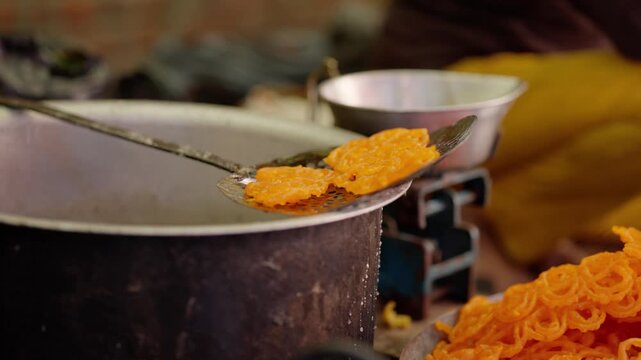 Perforated metal skimmer lifting hot jalebi sweets from a large pot, with another tray of orange spirals and blurred stove in background