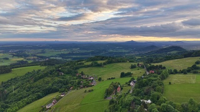 Panoramic dusk view over rolling green hills and patchwork fields, with farmhouses, winding lanes, and distant mountain ridges beneath dramatic clouds and soft sunset glow, conveying calm rural life.