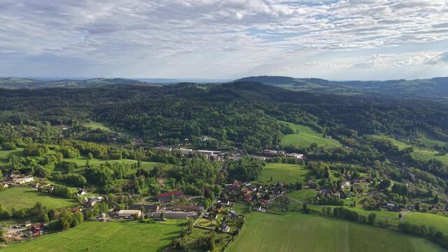 Wide aerial view of rural European landscape showing mosaic of fields, green meadows, scattered villages, and dense forests. Rolling hills rise toward horizon beneath sky with scattered clouds.
