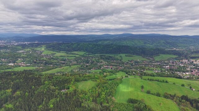 Sweeping overhead view shows quilted fields, forested slopes, and scattered buildings along winding roads, bordered by meadows and tree lines under stratified moody clouds.