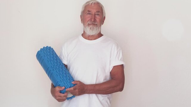 Fitness equipment store ad, senior man in white t-shirt with blue foam roller on light background. Home gym equipment for seniors and product demonstration, man turns roller upright and back