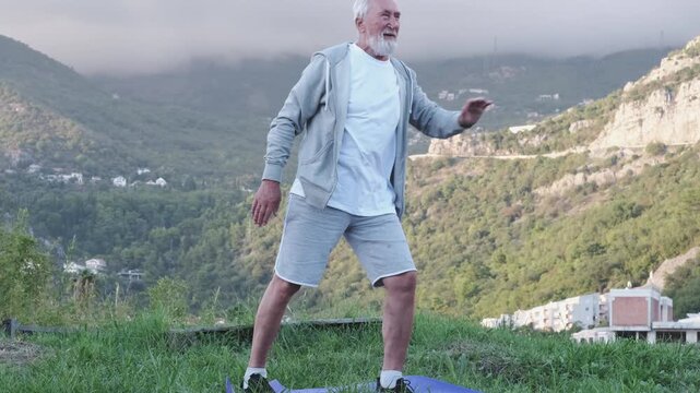 Balance training and fall prevention for elderly, senior man in shorts doing slow stretching pose on yoga mat on grass against mountains. Balance and tai chi ads, man holds pose with arm out