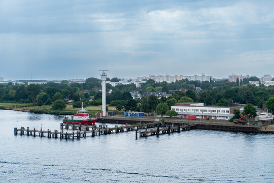 Coastal harbor scene with a tall navigation tower, red workboat, pier facilities and residential neighborhoods beyond a green waterfront. Maritime infrastructure and urban shoreline create a commercia