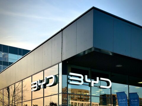A close-up photograph of a modern BYD car dealership building featuring prominent silver metallic BYD logo signage, glass facades, dark cladding panels, and a clear sky background.