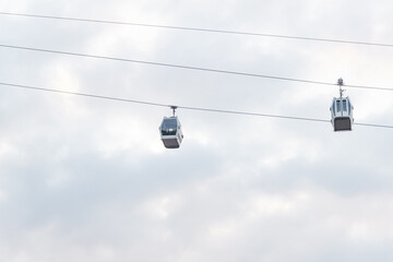 Two enclosed cable cars hang from parallel wires at different distances, silhouetted against a softly textured cloudy sky above © Anat art
