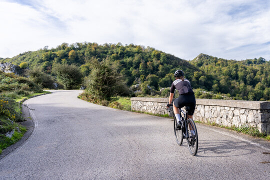 A female cyclist rides uphill along a winding road bordered by trees and a stone wall in Lagos de Covadonga, Picos de Europa, Spain. Viewed from behind, the scene captures the physical challenge of ro
