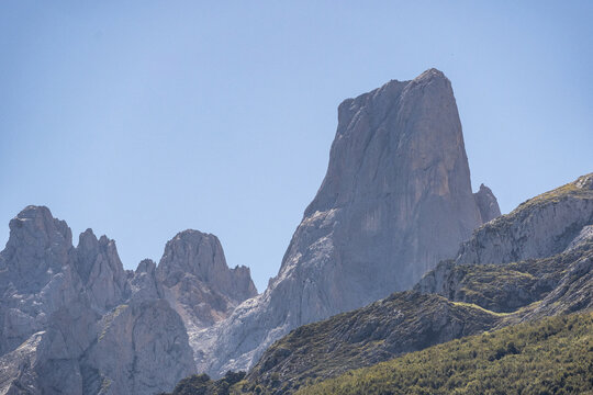 The steep rock face of Picu Urriellu, also known as Naranjo de Bulnes, rises above rugged slopes beneath a clear blue sky in Picos de Europa, Asturias, Spain.