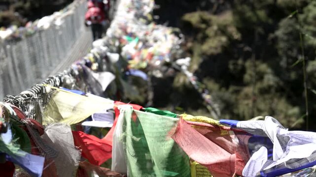 Traditional tibetan prayer flags fluttering in wind on a high suspension bridge in the himalayas, with a trekker walking across in the background over a deep green valley