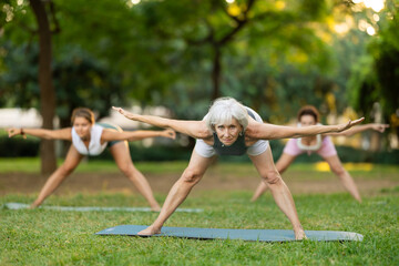 Fototapeta premium Motivated fit elderly woman participating in outdoor yoga session in green city park on summer day with group of female friends, enjoying healthy and active lifestyle