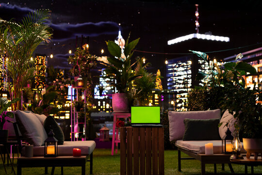 Isolated screen laptop on rooftop terrace table next to rose in stylish outdoor lounge; chroma key notebook among plants and cozy furniture with city.