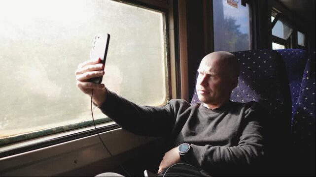 Smiling solo traveler takes selfie with smartphone beside train window and shares railway journey and vacation experience through social media while traveling.