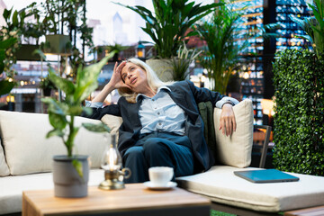 Fototapeta premium Fatigued businesswoman reclines with one hand on forehead during break, appearing overwhelmed with closed laptop beside her; relaxing on urban rooftop terrace.