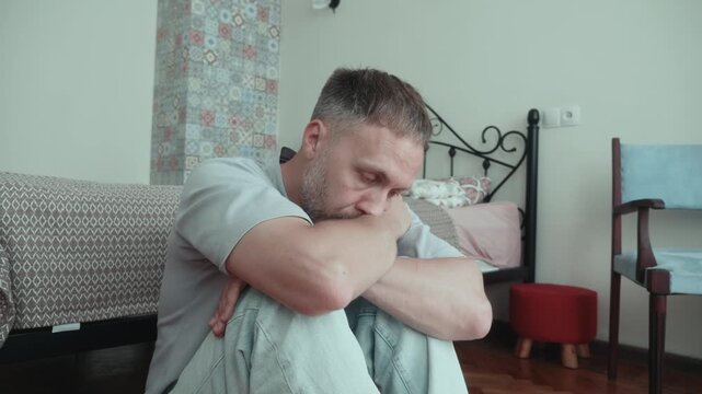 Man overwhelmed by burnout rests head on folded arms over knees at home, drained by hopelessness, low motivation, sadness and fatigue while sitting alone.