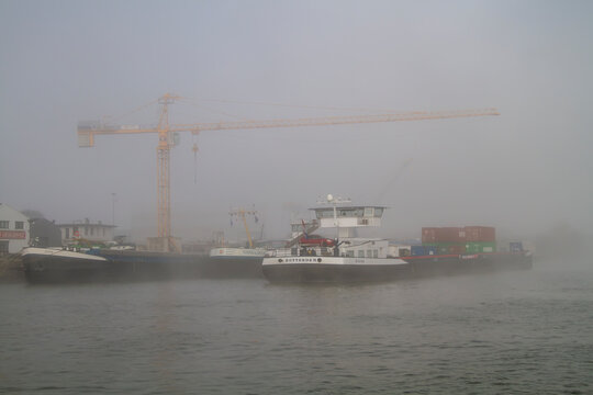 Rotterdam, Netherlands - March 11, 2015: Cargo vessels rest along the mist-covered Oude Maas near Rotterdam, framed by cranes and containers. Ideal for logistics, shipping, and industry concepts.