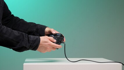 Person's hands holding a black wired video game controller while actively pressing buttons, representing gaming, leisure, and entertainment against a teal background © Aliaksandr